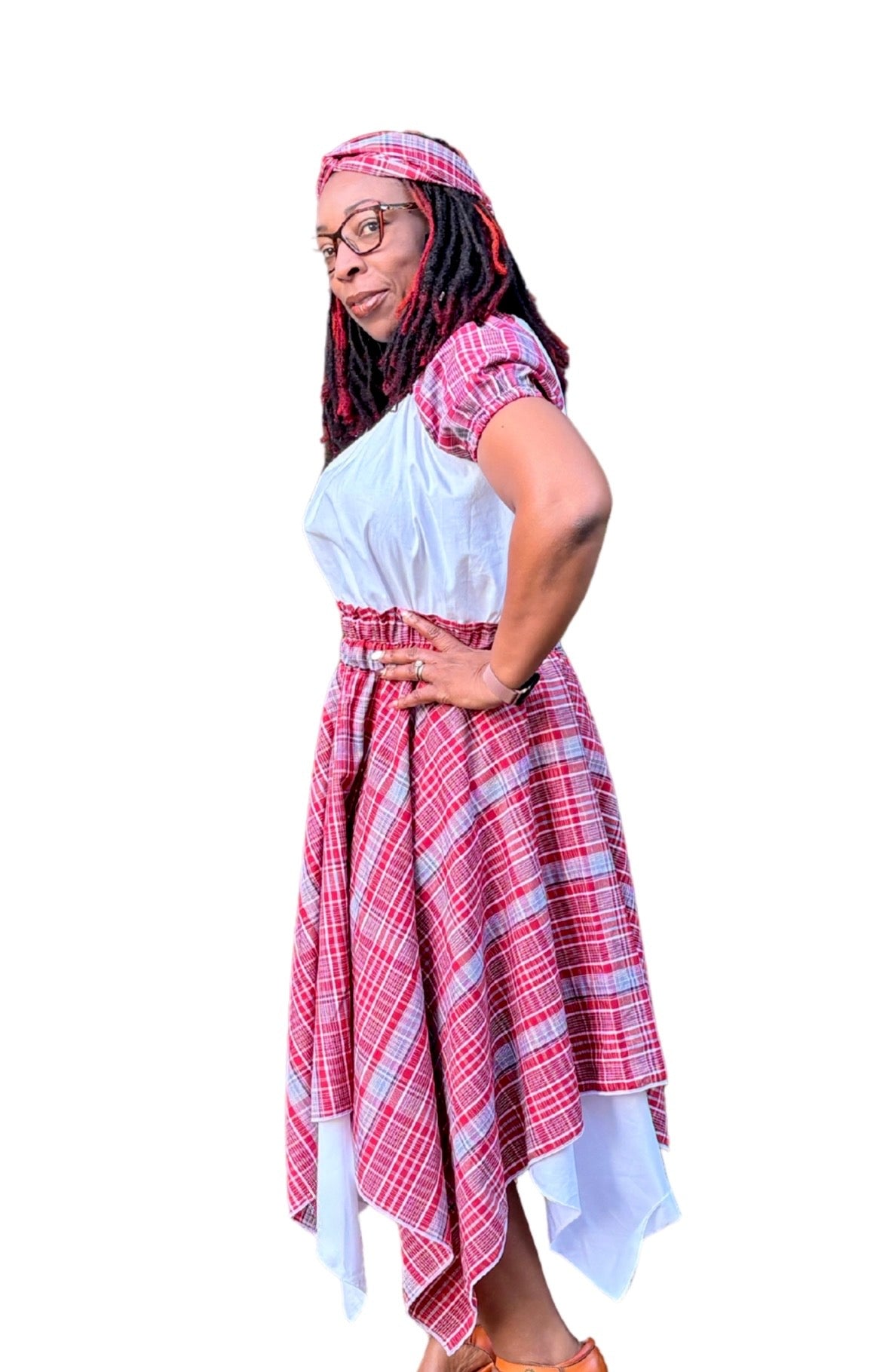 Side view of a Jamaican woman wearing a red, white, and blue bandana plaid skirt with asymmetrical hem over white underskirt, paired with a white peasant blouse with bandana sleeves and a matching twisted knot headband. Cultural outfit for International Day, Jamaica Day, or heritage events.