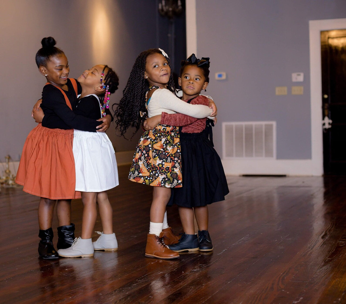 Four young girls hugging on a wooden floor in a room with a white wall and door.