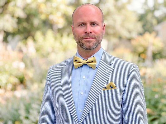 Man wearing a striped navy suit with a yellow with navy horses and navy dots design bow tie and pocket square