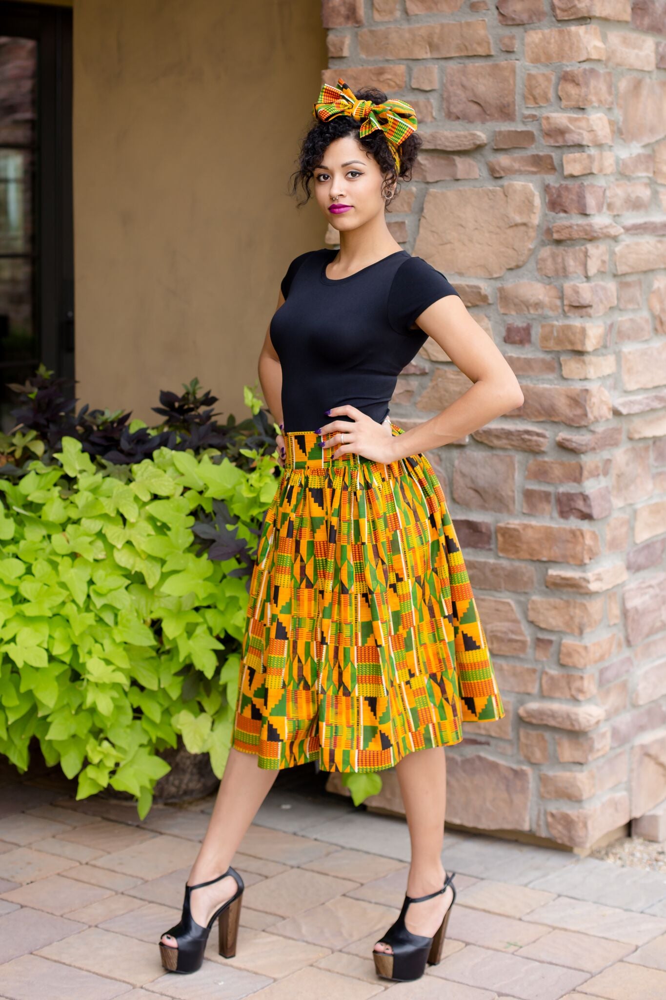 African American young woman in an African print Kente skirt and matching headwear and in a black tshirt and black heels. 