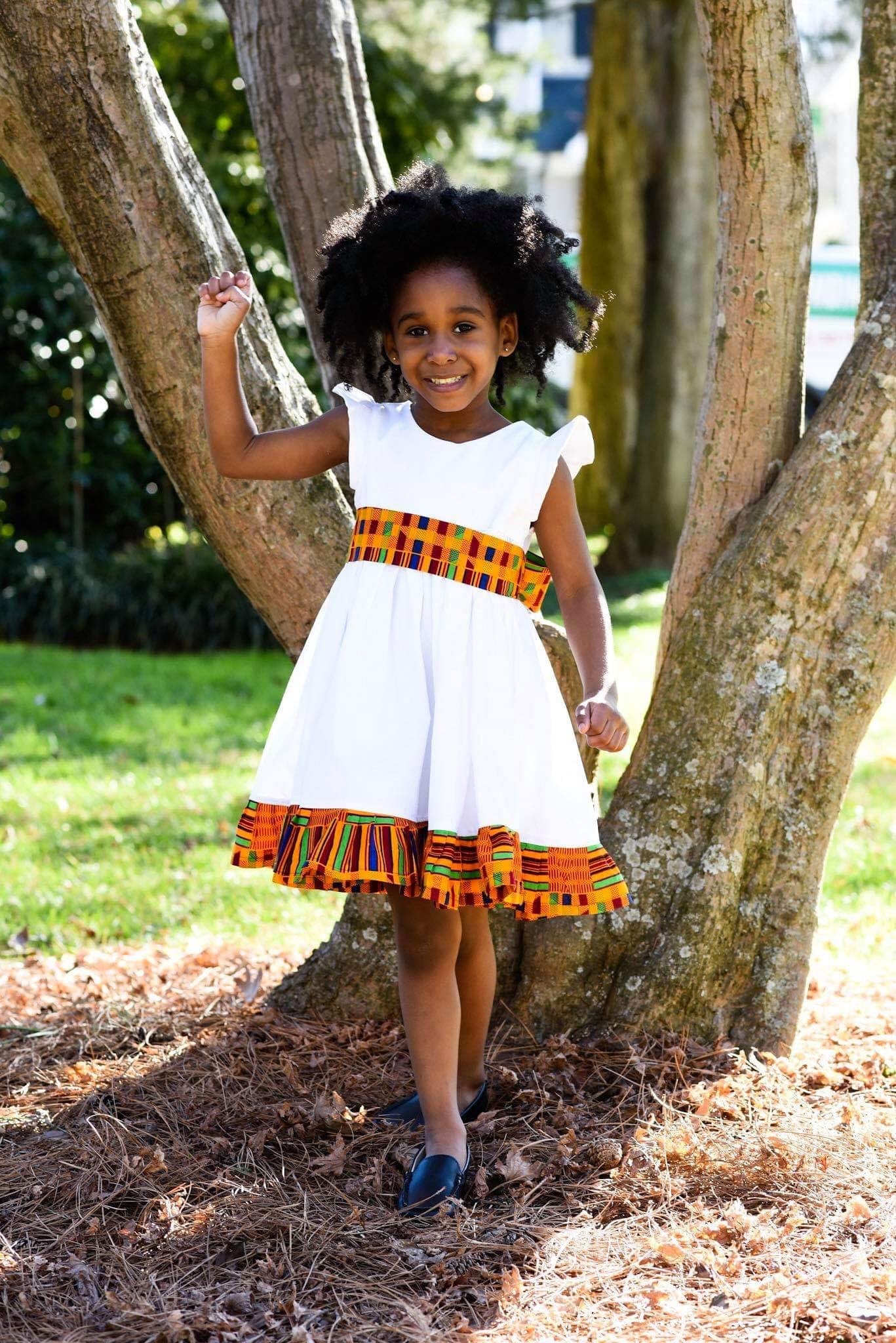 African American girl wearing white flutter sleeve dress with Orange Kente African print waist sash and ruffled hem.