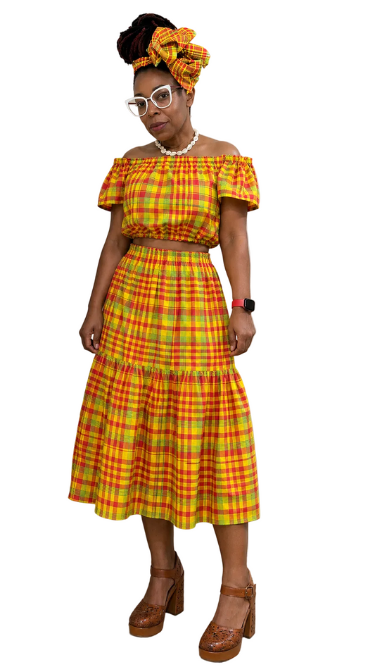 Woman wearing a yellow and red plaid madras Caribbean off the shoulder top and tiered skirt outfit with a head wrap, standing against a white backdrop.