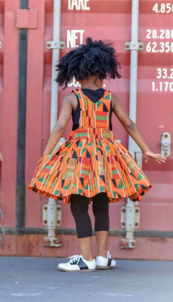 Back view of African American girl wearing orange Kente pinafore dress.