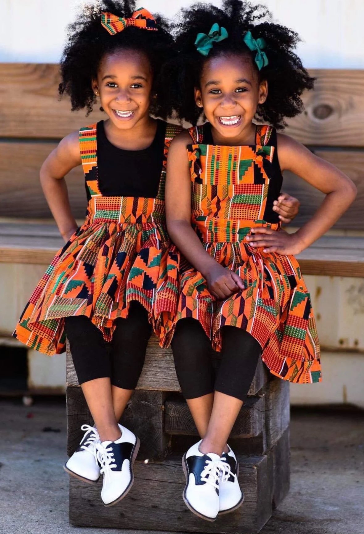 African American Twin girls hugging and wearing orange Kente suspender skirt and a pinafore dress.