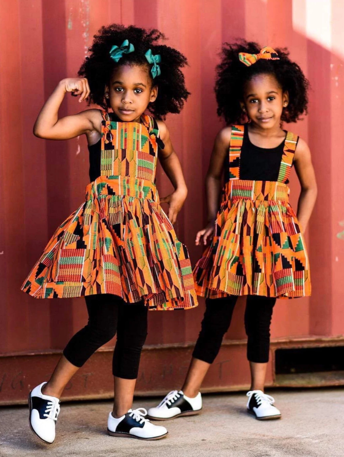 African American Twin girls wearing orange Kente suspender skirt and a pinafore dress with hair bow