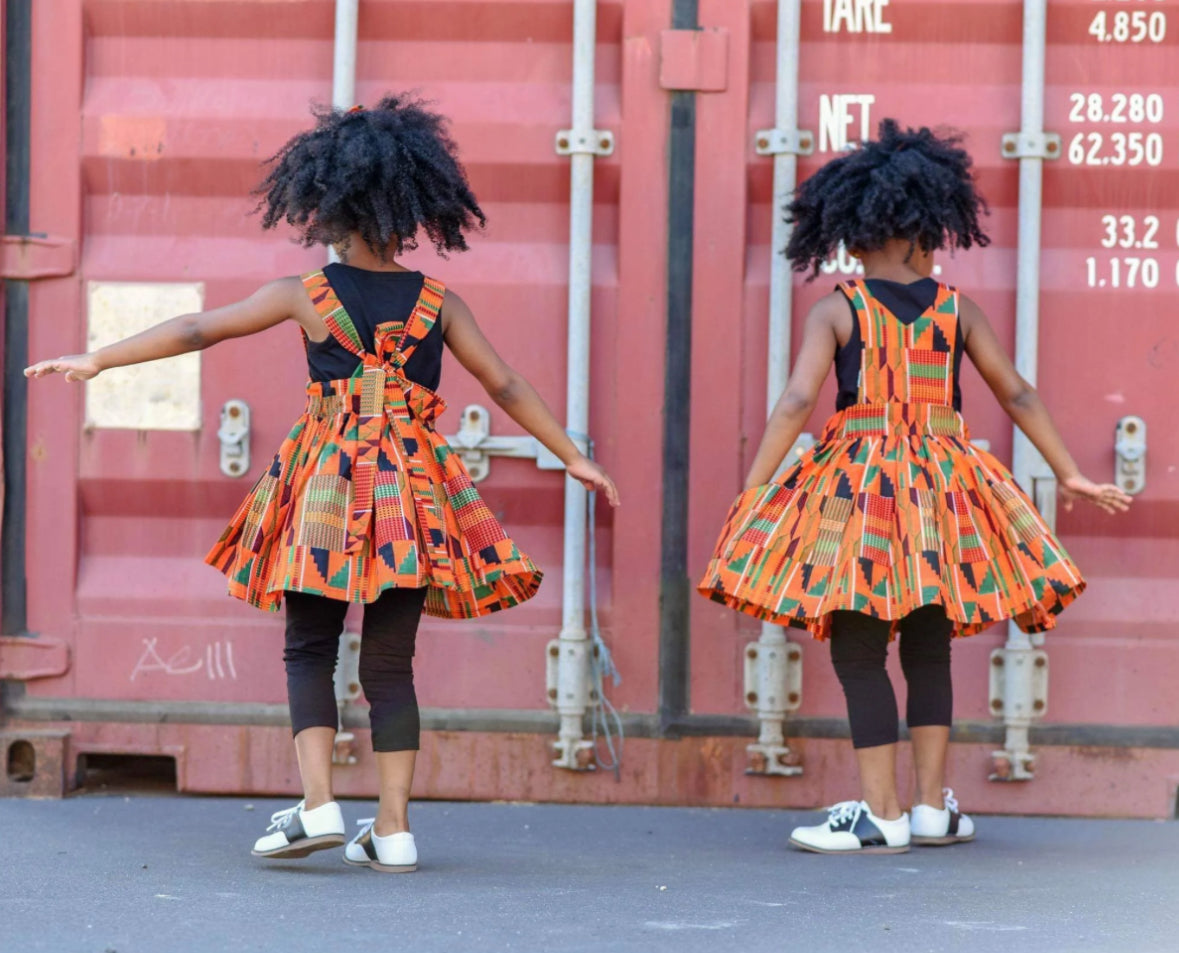 Back view of African American Twin girls wearing orange Kente suspender skirt and a pinafore dress.