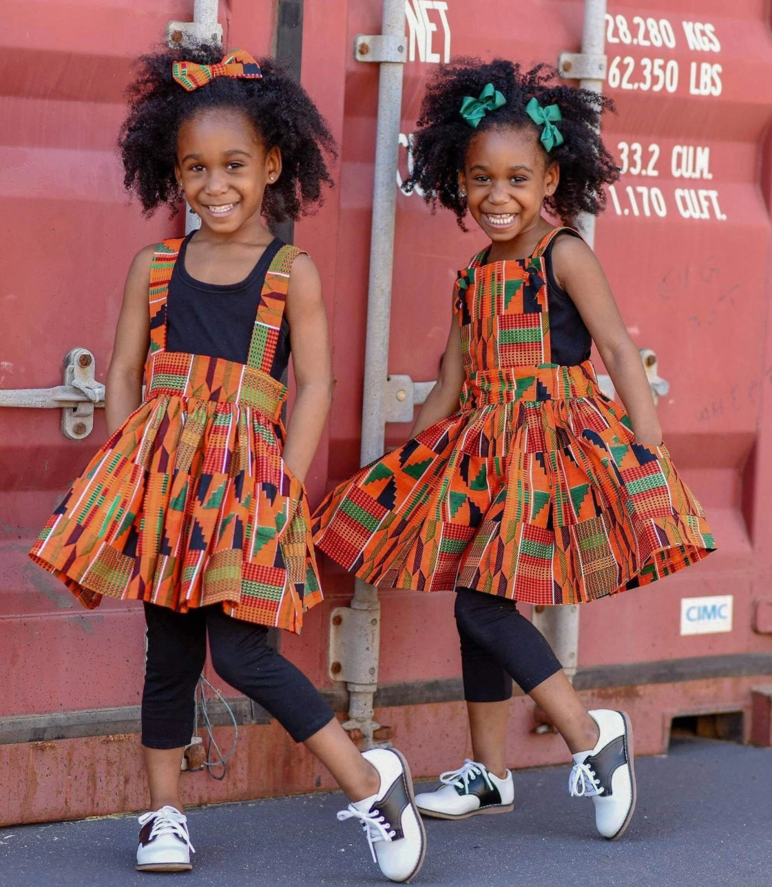 African American Twin girls wearing orange Kente suspender skirt and a pinafore dress.