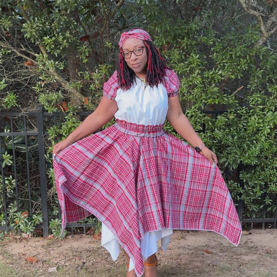Video of a Jamaican woman wearing a red, white, and blue bandana plaid skirt with asymmetrical hem over white underskirt, paired with a white peasant blouse with bandana sleeves and a matching twisted knot headband. Cultural outfit for International Day, Jamaica Day, or heritage events.