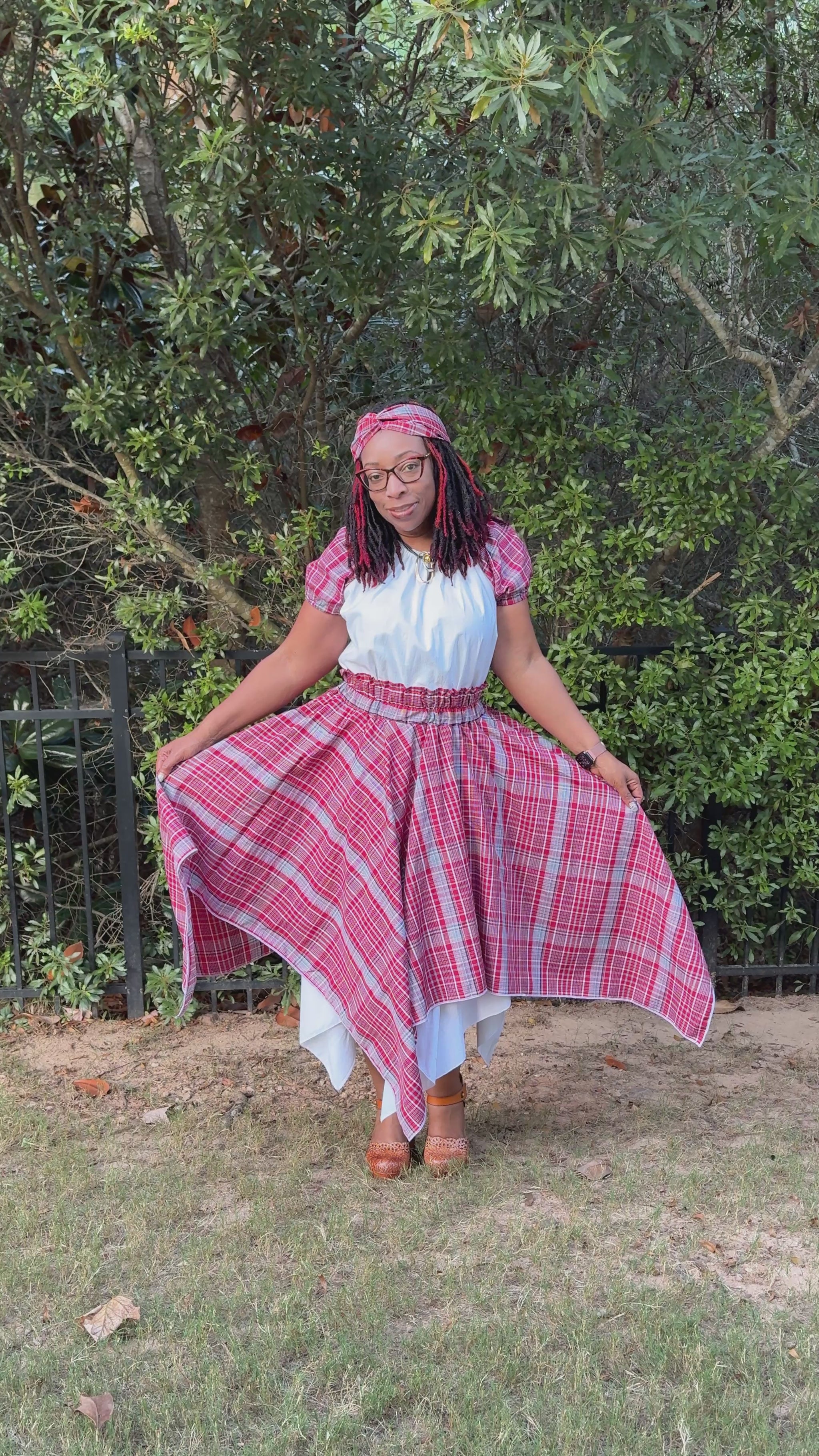 Video of a Jamaican woman wearing a red, white, and blue bandana plaid skirt with asymmetrical hem over white underskirt, paired with a white peasant blouse with bandana sleeves and a matching twisted knot headband. Cultural outfit for International Day, Jamaica Day, or heritage events.