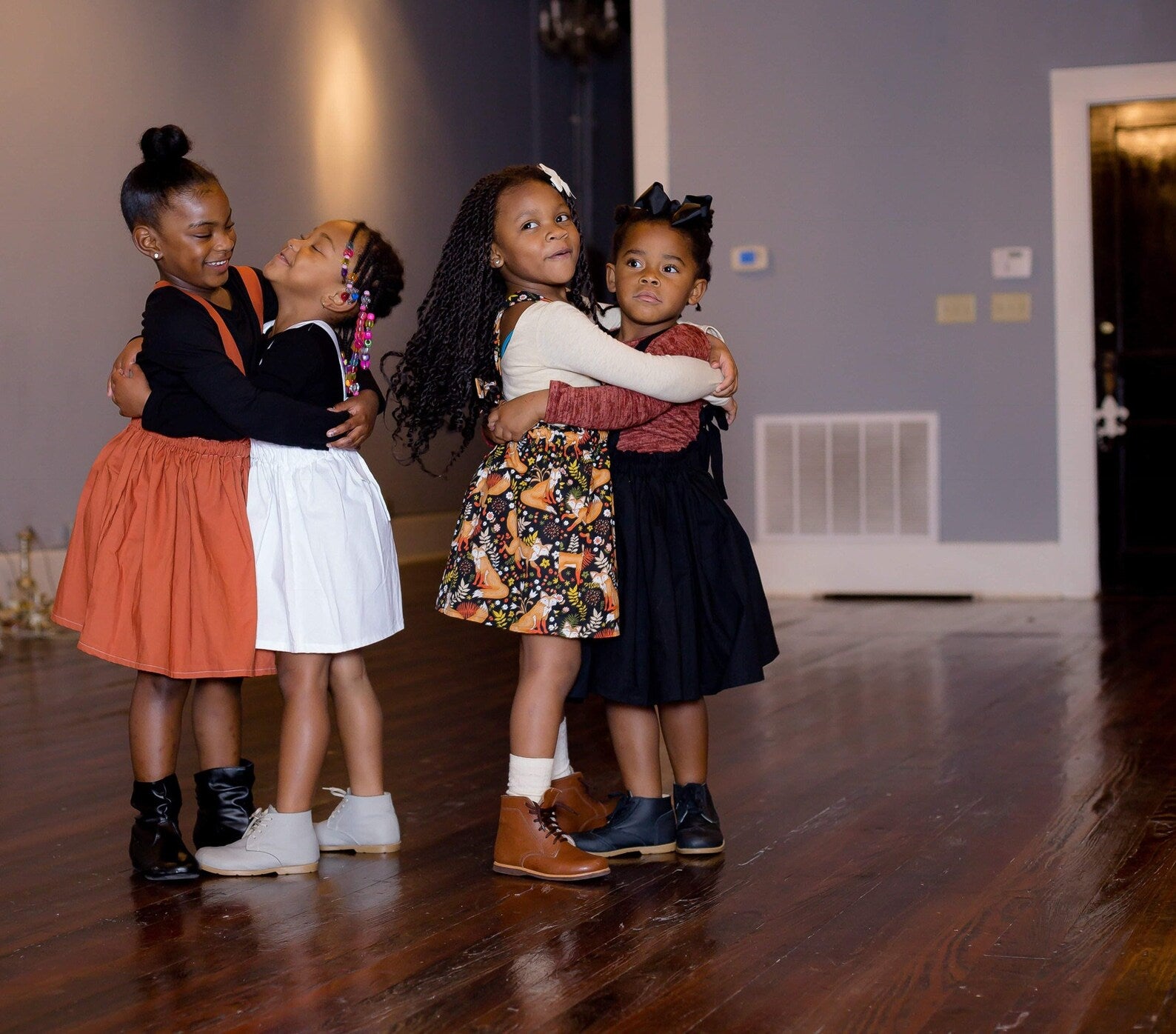 Four young girls hugging on a wooden floor in a room with a white wall and door.
