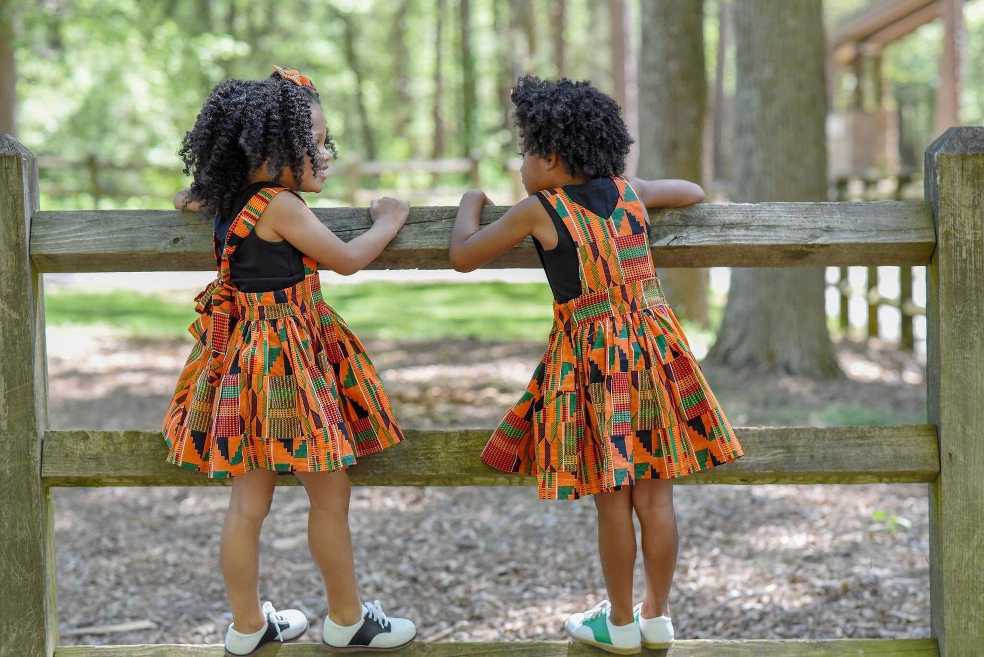 Two African American girls playing on a fence wearing orange Kente suspender skirt and a pinafore dress.