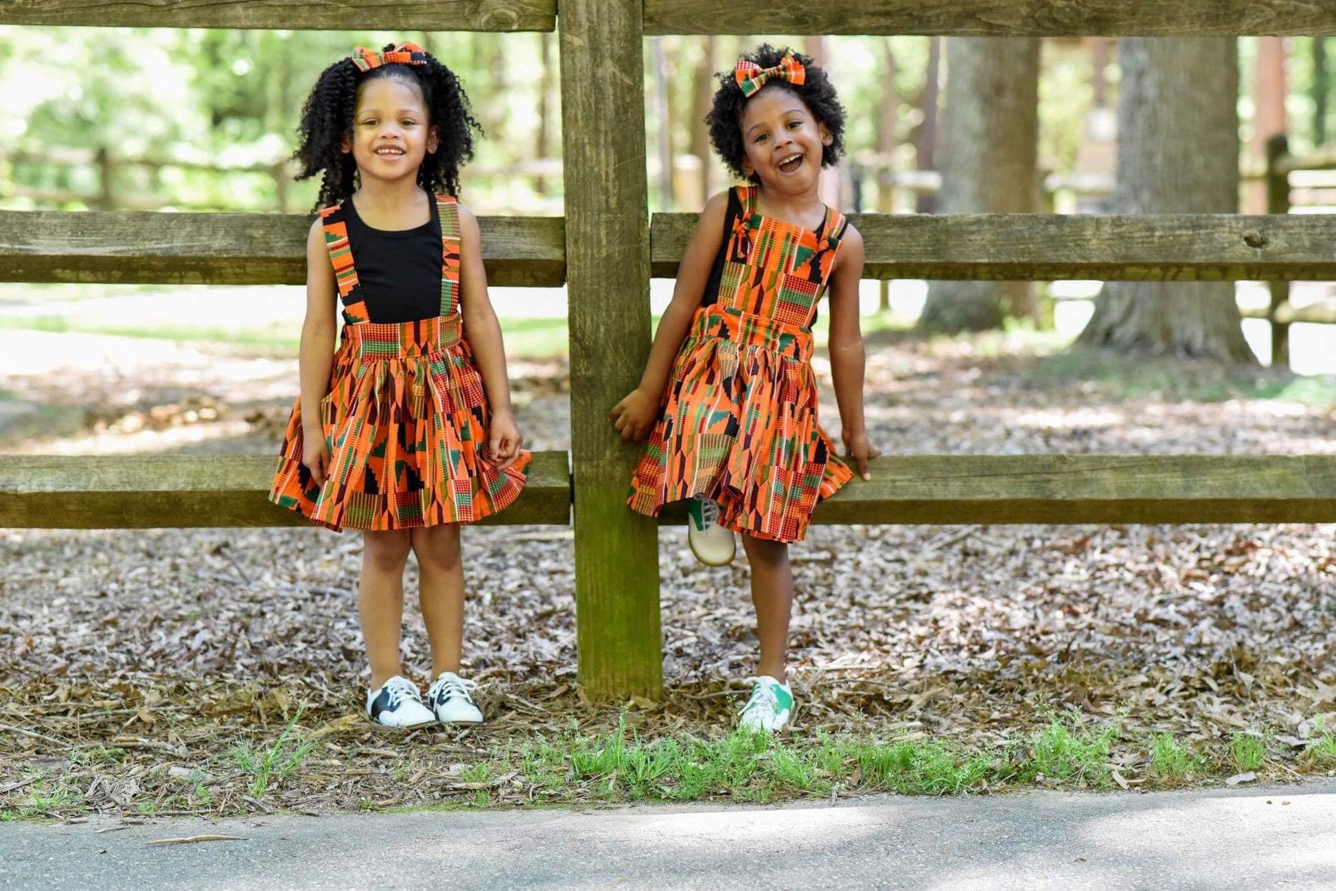 African American girls posing in coordinating African print orange Kente suspender skirt and pinafore dress with hair bows.