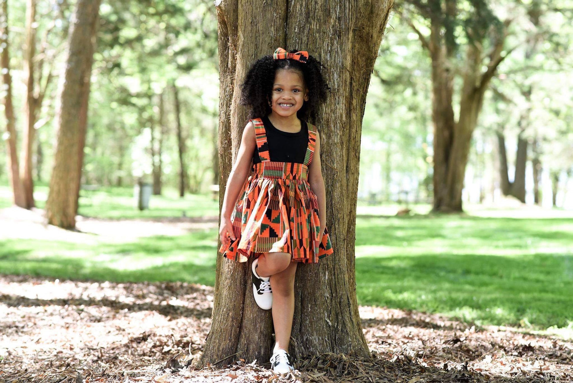 African American girl wearing orange Kente suspender skirt and a matching hairbow.