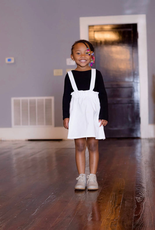 A young girl wearing a white suspender skirt with a black top, standing in a home environment.